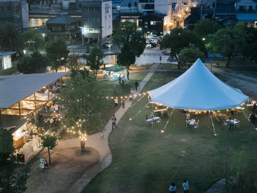 夕暮れの福山中央公園を見下ろした様子。芝生広場の大型テントに灯りがともり、園内を人が行き交っている。奥には福山のまちなみが広がる。