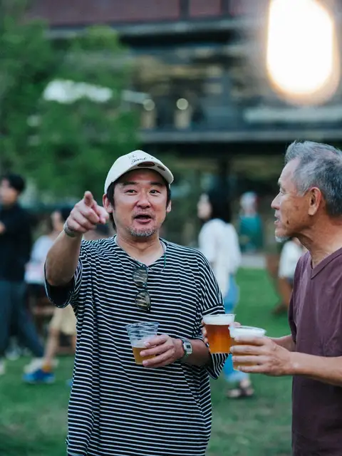 夕暮れの芝生の広場で、生ビールのカップを手に笑顔で語らう2人の来場者。