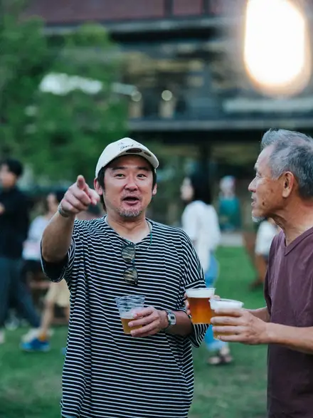 夕暮れの芝生の広場で、生ビールのカップを手に笑顔で語らう2人の来場者。