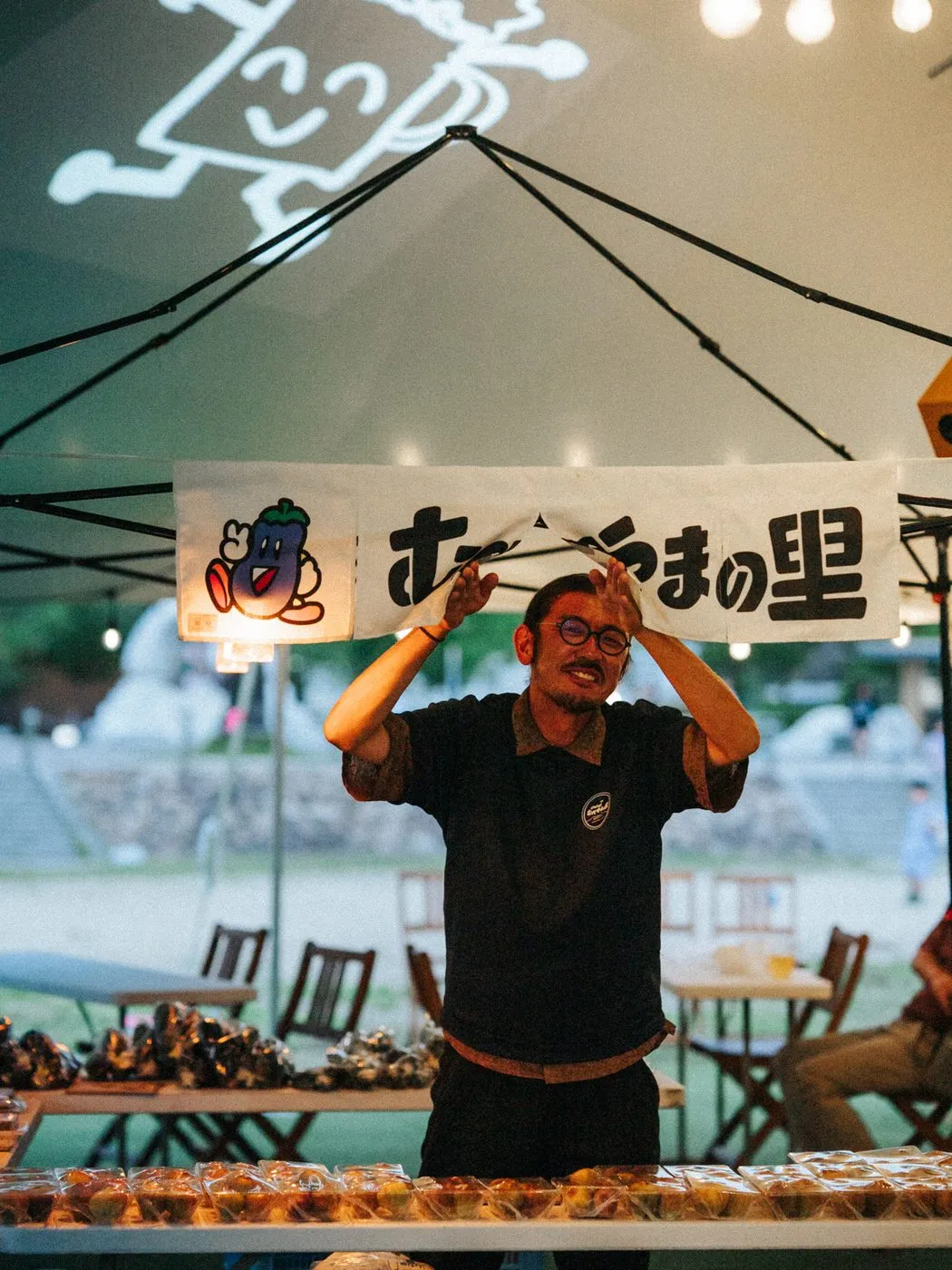 「むべやまの里」ののれんの前で、両手を上げてポーズをとる店主。テーブルには旬の野菜と加工品が並ぶ。